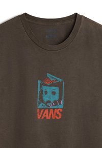 Tmavě hnědé tričko vyrobené z bavlny, na přední straně s barevným grafickým motivem obličeje panenky a textem "VANS" v červené barvě. Klasický střih s kulatým výstřihem.