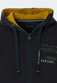 Black zip-up hoodie with a yellow lining, drawstrings, and graphic text details. Features durable fabric and a modern design.
