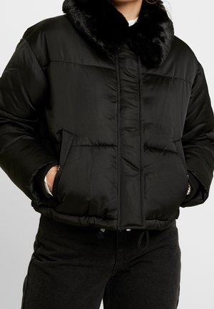 Person wearing a black puffer jacket with a soft fur collar, hands in front pockets, paired with black pants.