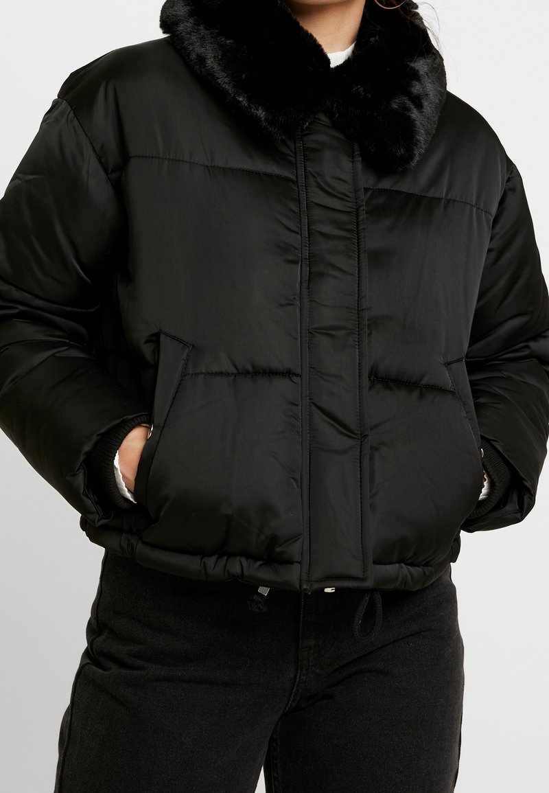 Personne portant une doudoune noire avec un col en fourrure douce, les mains dans les poches avant, associée à un pantalon noir.