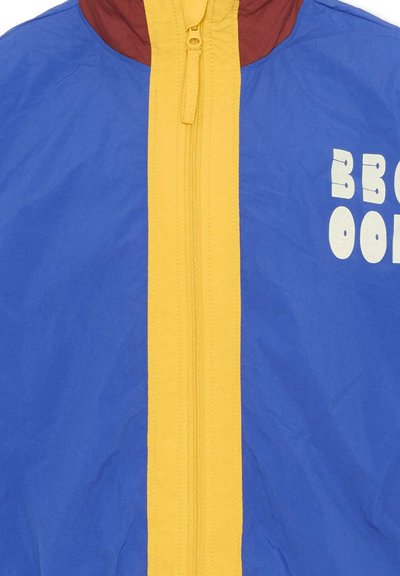 Veste bleue avec une fermeture éclair centrale jaune, col bordeaux, et texte blanc partiel sur le côté droit indiquant "BRO" et "OO".