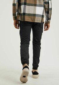 Persoon in zwarte joggingbroek, beige en zwarte sneakers, en een bruine, witte en zwarte geruite blouse, die wegloopt op een effen achtergrond.