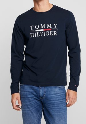 Hombre con camisa de manga larga azul marino de Tommy Hilfiger con logo y bloque de color encima del texto, combinado con jeans azules.