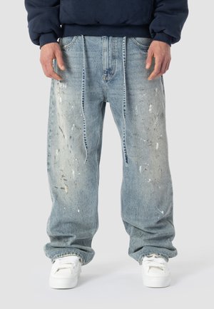 Osoba nosząca luźne, jasnoniebieskie dżinsy z plamami farby i białe trampki, stojąca na tle jednolitego szarego tła.