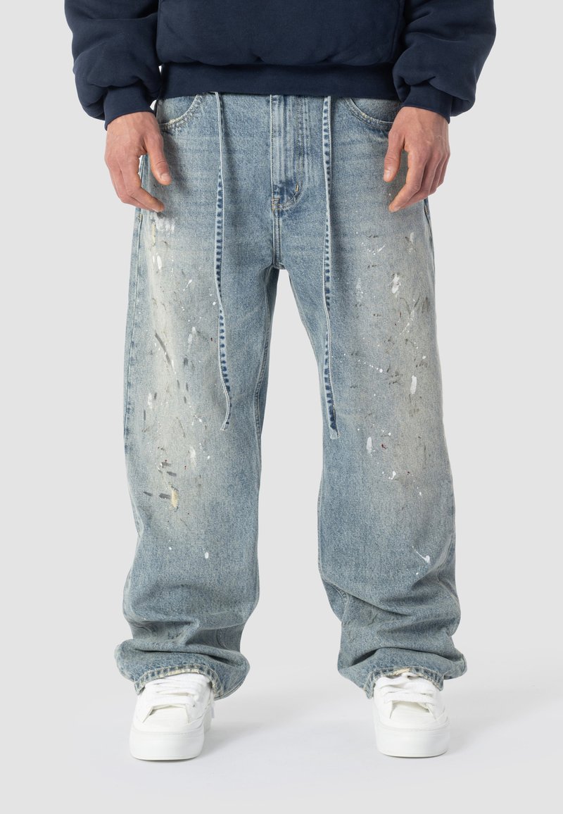 Persona che indossa jeans in denim azzurro chiaro, larghi e con schizzi di vernice, e scarpe da ginnastica bianche, in piedi contro uno sfondo grigio uniforme.