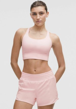 Femme debout, les mains derrière le dos, portant un soutien-gorge de sport rose clair et un short de sport assorti sur un fond uni.