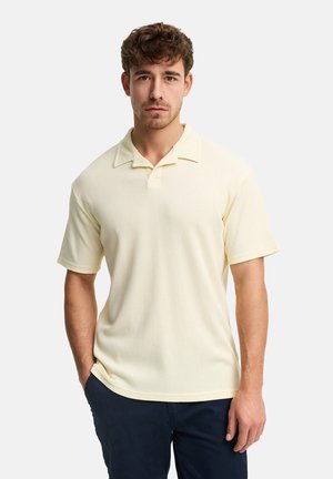 Junger Mann mit lockigem Haar, der ein cremefarbenes, kurzärmeliges, strukturiertes Poloshirt und dunkle Hosen trägt, mit einer Hand in der Hosentasche stehend.