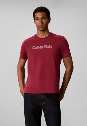 Homme portant un t-shirt rouge à manches courtes avec "Calvin Klein" imprimé sur la poitrine et un jean foncé, debout devant un fond gris.
