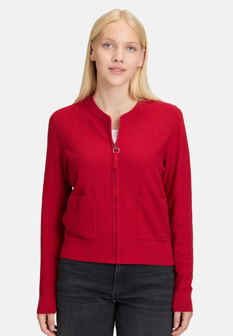 Czerwona kardigan zapinany na zamek, wykonany z teksturowanego materiału dzianinowego, z okrągłym dekoltem, dwiema przednimi kieszeniami i ściągaczami przy rękawach.