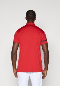 Camisola polo vermelha com colarinho e mangas curtas, apresentando detalhes pretos nas mangas. Feita de um tecido leve e suave.