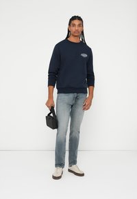 Felpa blu navy con collo tondo, polsini a coste e un logo. Abbinata a jeans azzurri chiari e sneakers crema, tiene una piccola borsa nera.