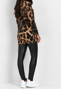 Femme portant un long pull marron et noir à imprimé léopard, des leggings en similicuir noir et des baskets blanches épaisses, debout de profil.