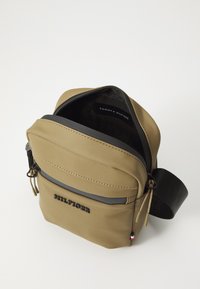 Geantă crossbody de culoare bej, realizată din material sintetic neted, având un buzunar frontal cu fermoar, curea ajustabilă și detalii cu logo pe față.