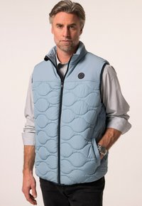 Světle modrá vyšívaná vesta s předním zipem a kulatou nášivkou. Nositá přes šedou košili s dlouhým rukávem, s tmavými džíny a hodinkami.