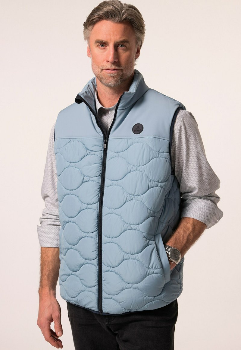 Světle modrá vyšívaná vesta s předním zipem a kulatou nášivkou. Nositá přes šedou košili s dlouhým rukávem, s tmavými džíny a hodinkami.