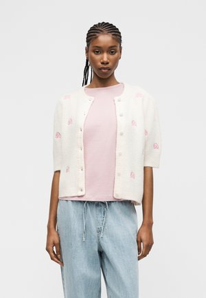 Fonott hajú nő fehér, pink mintás, kigombolt kardigánt visel világos rózsaszín ing és világoskék farmer felett.