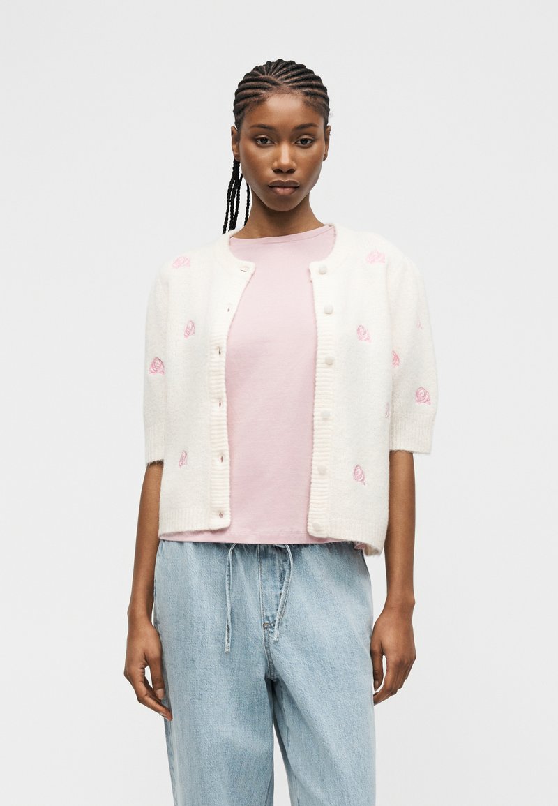 Γυναίκα με πλεξούδες, φορώντας ένα λευκό ζακέτα με κουμπιά και ροζ σχέδια πάνω από ένα ανοιχτό ροζ πουλόβερ και ανοιχτό μπλε τζιν.