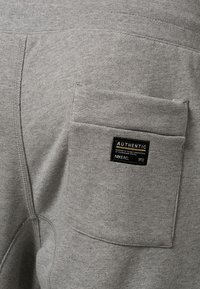 Grå sweatshirt-tyg med en sidoficka som har en svart etikett med texten "AUTENTISK, NIKE FC, 1972." Texturerad sömdetalj synlig.