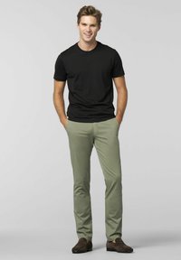 Zwarte crew-neck t-shirt gecombineerd met lichtgroene slim-fit broek en bruine instap schoenen. Model staat met handen in de zakken, glimlachend.