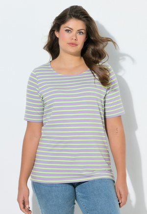 Kobieta o długich brązowych włosach ubrana w fioletową koszulkę z krótkim rękawem w żółte paski oraz niebieskie dżinsy, stoi na białym tle.