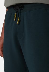Pantalones de chándal en color verde azulado oscuro con cintura ajustable mediante un cordón negro y dispositivos de ajuste en color verde oliva. Tejido suave y detalle de bolsillo lateral.