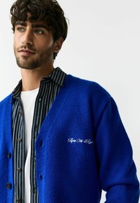 Blauwe gebreide cardigan met een gestructureerde afwerking, voorzien van een V-hals, knopen aan de voorkant en een geborduurd logo op de borst. Draag over een gestreept shirt.