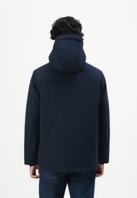 Mann iført en marineblå hettejakke som snur ryggen til mot en enkel hvit bakgrunn.