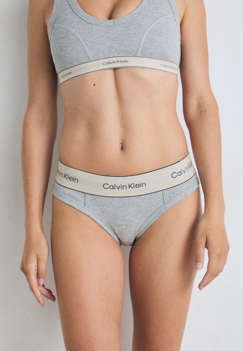 Szürke pamut sportmelltartó bézs Calvin Klein logós pánttal, amelyet a hozzá illő, szürke, magas derekú bikini alsóval kombináltak, szintén ugyanazzal a logóval.