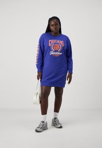 Μωβ φόρεμα-παντελόνα με εκτυπώσεις γραφικών στο μπροστινό μέρος και στα μανίκια; διαθέτει στρογγυλή λαιμόκοψη, μακριά μανίκια και άνετη εφαρμογή. Συνδυάζεται με αθλητικά παπούτσια και τσάντα.