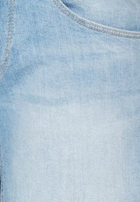 Tela de mezclilla azul claro, con una textura suave y variaciones sutiles. Incluye un diseño de bolsillo estándar y detalles de costura en contraste.