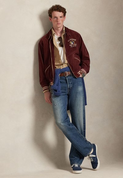 Bordo manšestarska jakna s kremastim detaljima, bež košulja, plavi džemper vezan oko struka, plave traperice, mornarskoplave tenisice s bijelim detaljima.