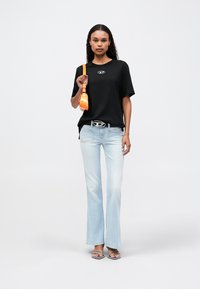 Camiseta negra de gran tamaño con un logo en el centro, jeans acampanados de lavado claro, bolso naranja y sandalias de tiras transparentes sobre un fondo liso.