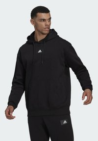 Sudadera negra de material polar con bolsillo canguro y capucha con cordón, que presenta un pequeño logo blanco de Adidas en el pecho.