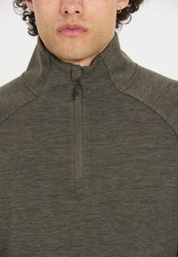 Olivengrøn zip-up pullover lavet af blødt, tekstureret stof. Har en høj krave og en kort lynlås ved halsen.