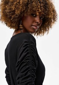 Femme avec des cheveux bouclés volumineux portant de grandes boucles d'oreilles en forme de anneau et un haut noir froncé à manches longues, regardant par-dessus son épaule.