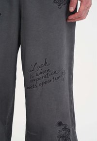 Szare bawełniane spodnie z czarnym haftowanym napisem i wzorem smoka na nodze. Gładka tekstura i luźny krój.