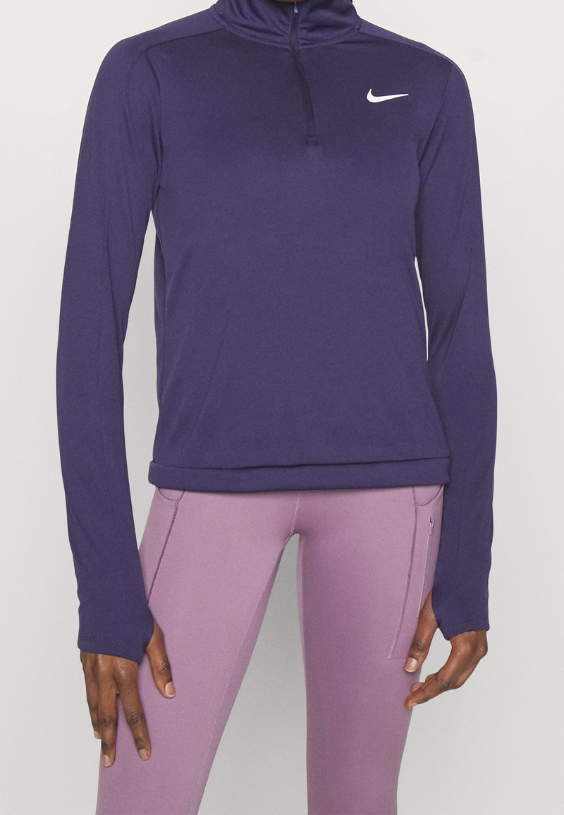 Person som bär en lila långärmad Nike-pullover med dragkedja och ljuslila leggings med sidoficka, står mot en enfärgad bakgrund.