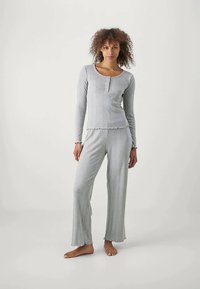 Femme debout pieds nus, portant un haut à manches longues gris clair avec des boutons et un pantalon large assorti, devant un fond uni.