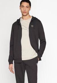 Sweat à capuche zippé gris foncé avec cordons, arborant un petit logo sur la poitrine. Porté par-dessus un t-shirt beige clair avec un design minimaliste en lignes.