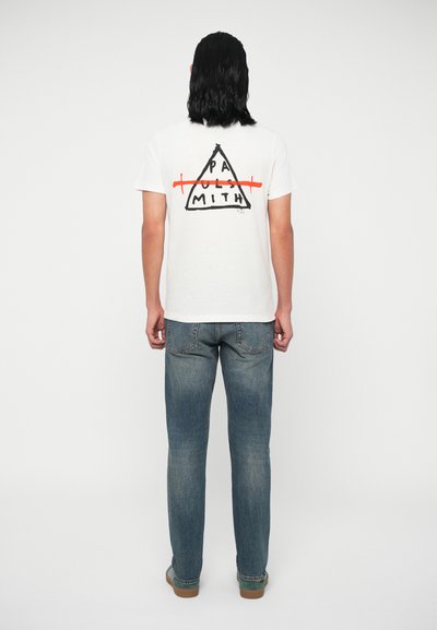 T-shirt blanc avec un graphique triangulaire au dos, comportant du texte noir "PA...SMITH" barré en orange. Assorti à un jean bleu.