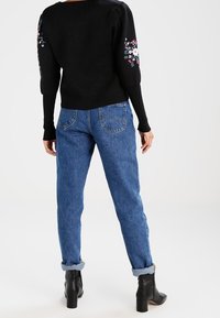 Pull noir avec broderie florale sur les manches, associé à un jean bleu clair retroussé et des bottines noires. Textures de tricot lisse et de denim.