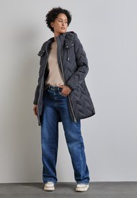 Quiltad mörkgrå jacka med dragkedjesl closure, justerbar huva och kontrasterande dragkedjedetaljer, i kombination med blå jeans och krämfärgade sneakers.