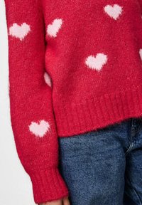 Czerwony sweter z dzianiny z białymi wzorami serc, noszony z niebieskimi dżinsami, pokazującym środkową część ciała i lewą rękę.