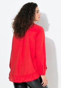Rode blouse met een gerimpelde zoom, lange mouwen, knoopdetails en een losse pasvorm. De stof lijkt glad met een lichte glans.