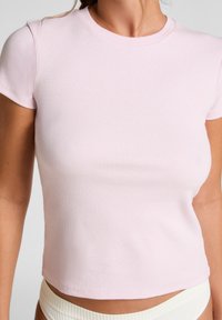 Vrouw in een lichtroze, korte mouw, geribbelde crop top en witte geribbelde onderkleding, getoond van nek tot heupen tegen een effen achtergrond.