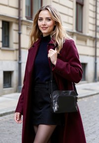 Borsa a tracolla in pelle nera con una texture liscia e finitura lucida, dotata di accessori in argento e un charm decorativo. Indossata su una tracolla.