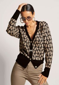 Der Cardigan verfügt über ein geometrisches Muster in Schwarz und Beige, gerippte schwarze Bündchen und einen Saum sowie Vorderknöpfe. Er wird mit hellen Hosen getragen.