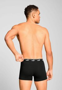 Muscular man seen from behind adjusting black Puma boxer briefs with branded waistband against plain light background.