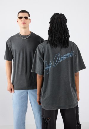 Zwei Personen tragen übergroße T-Shirts in dunkelgrau mit blauer Aufschrift "Only Dreamers" auf dem Rücken. Der Stoff wirkt weich und hat einen lässigen Schnitt.