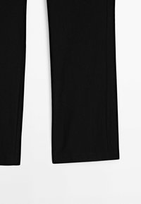 Partie inférieure d’un pantalon noir à jambes droites posé à plat sur une surface blanche, montrant la texture du tissu et les détails de l’ourlet.